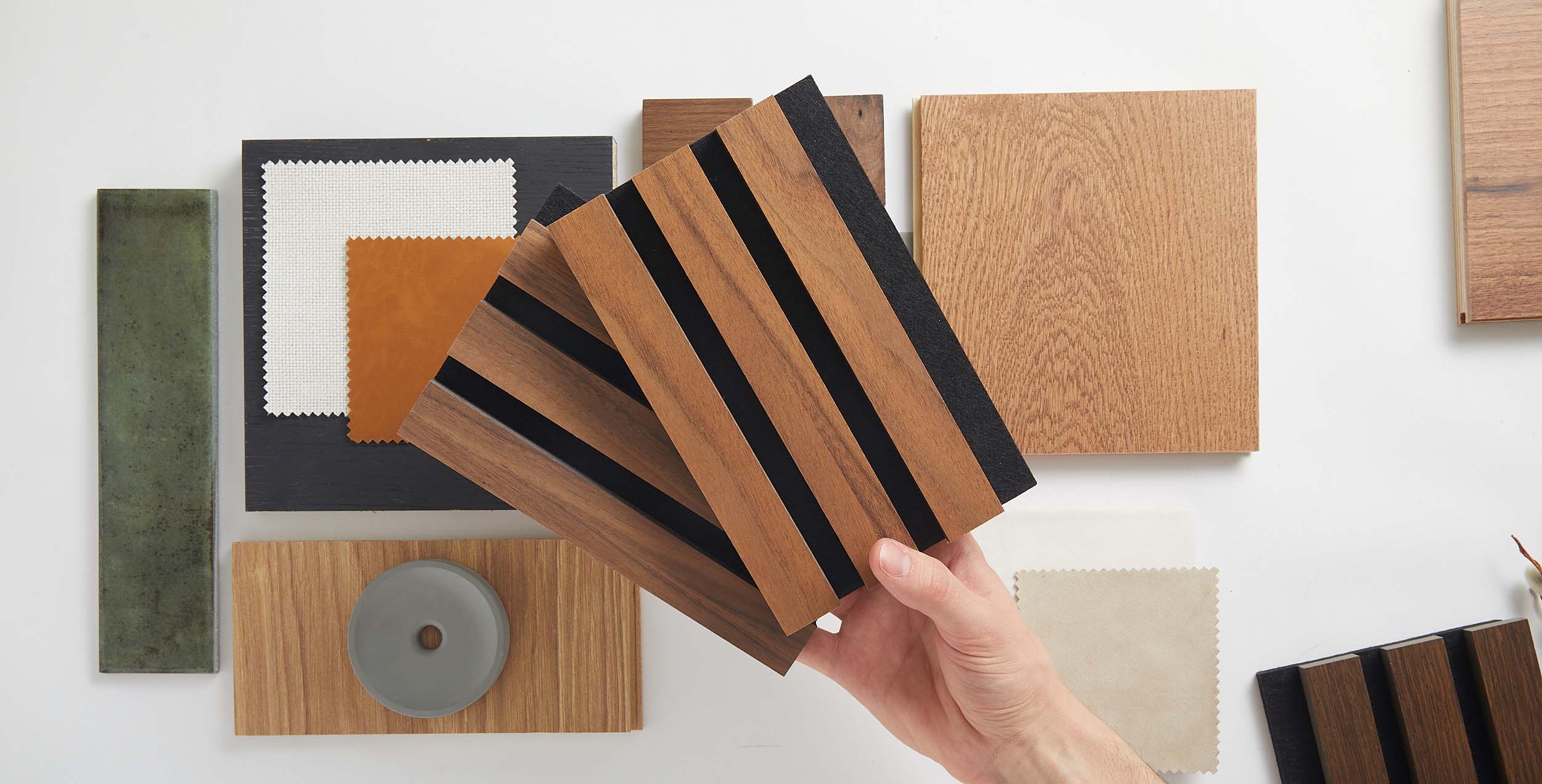 A person's hand holding a sample of dark wood slat wall panels, surrounded by various material swatches and textures for interior design.