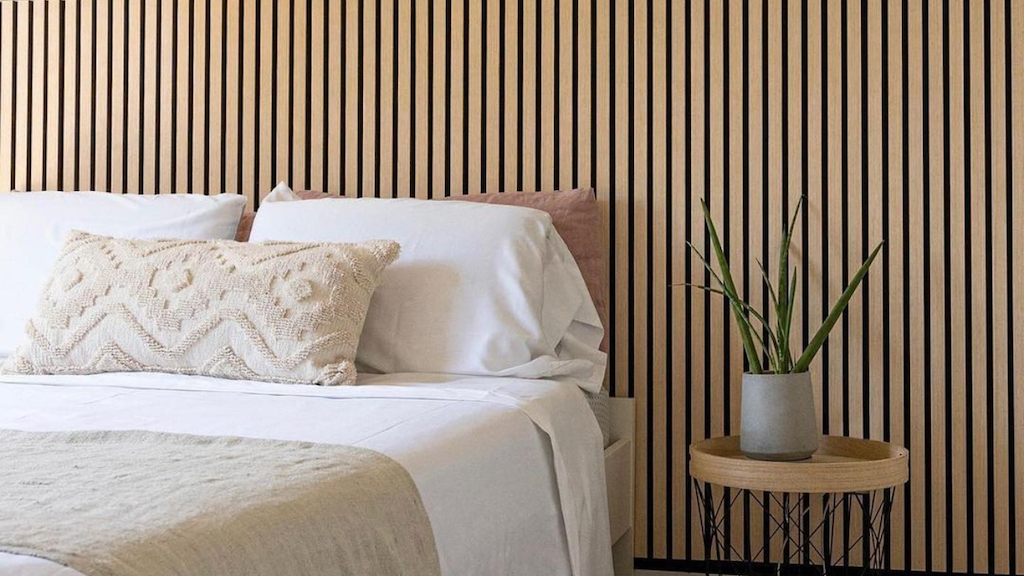 A minimalist home bedroom featuring a bed with white linens, a textured beige pillow, and a wooden slat wall paneling backdrop, alongside a small round side table with a potted plant.