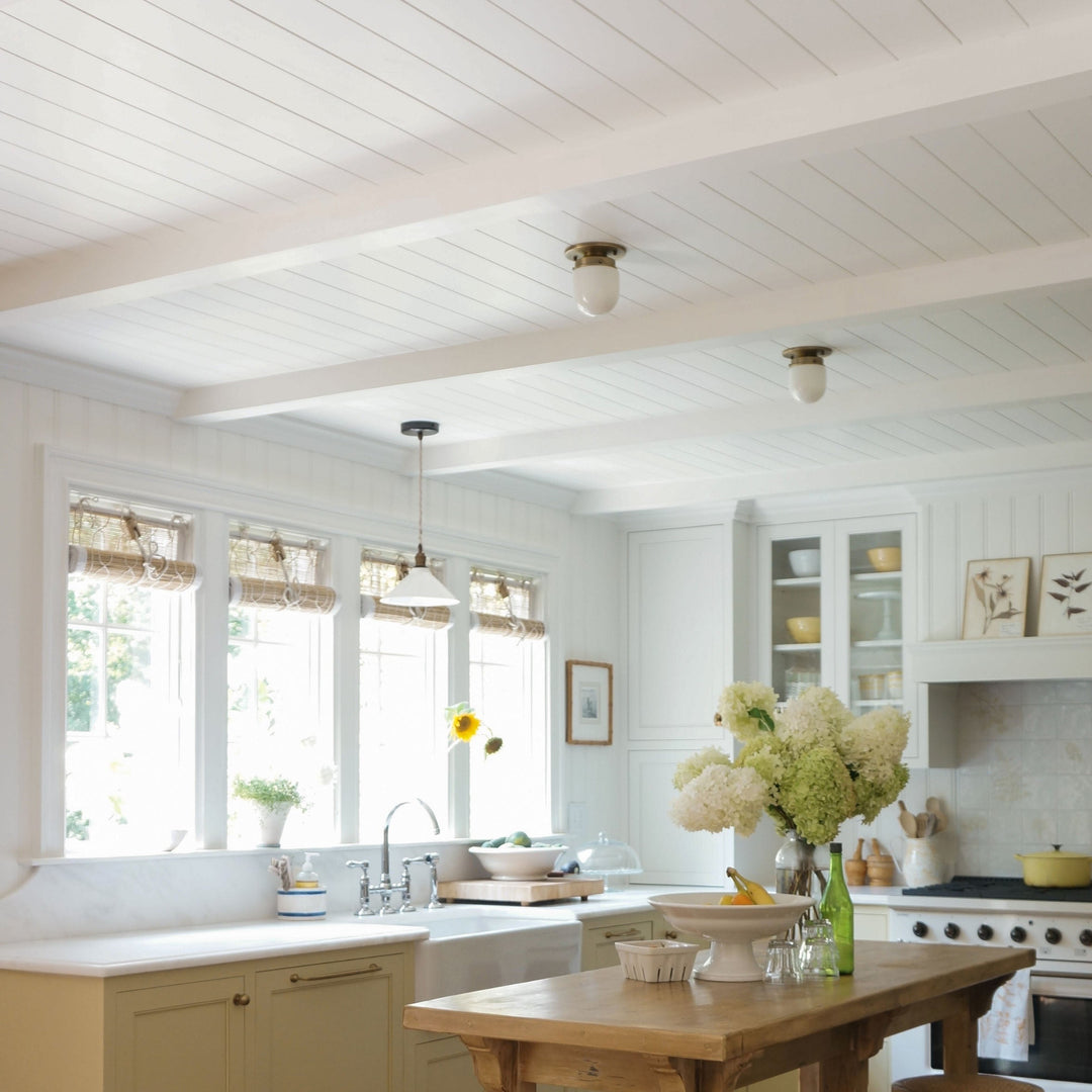Bright, airy kitchen interior with white shiplap ceiling and walls, cream cabinets, a wooden island, large windows with woven shades, and a vase of white flowers on the counter.