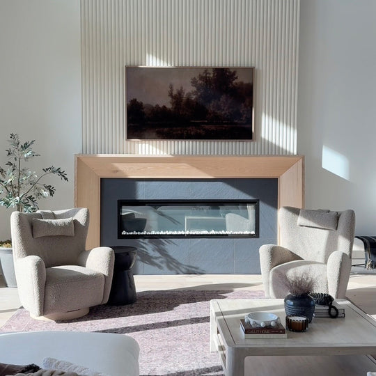 A modern living room featuring a built-in linear fireplace with a light wood mantel, flanked by two textured beige armchairs. A large framed landscape painting hangs on the ribbed white wall above the fireplace.