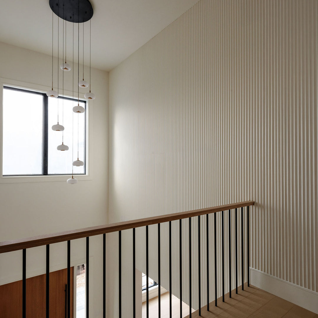 Modern interior entryway with fluted white wall paneling, wooden handrail, black metal balustrade, and a multi-light pendant chandelier.