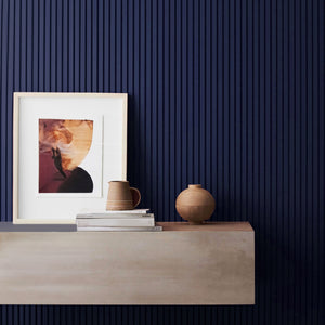 A modern living room corner with a slatted paintable wall panel in deep blue, adorned with a framed artwork, books, and rustic pottery on a wooden console table.